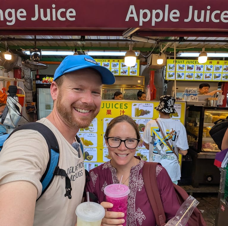 Don and Samantha holding fresh fruit juices and snacks at a vibrant Asian street food market stall.