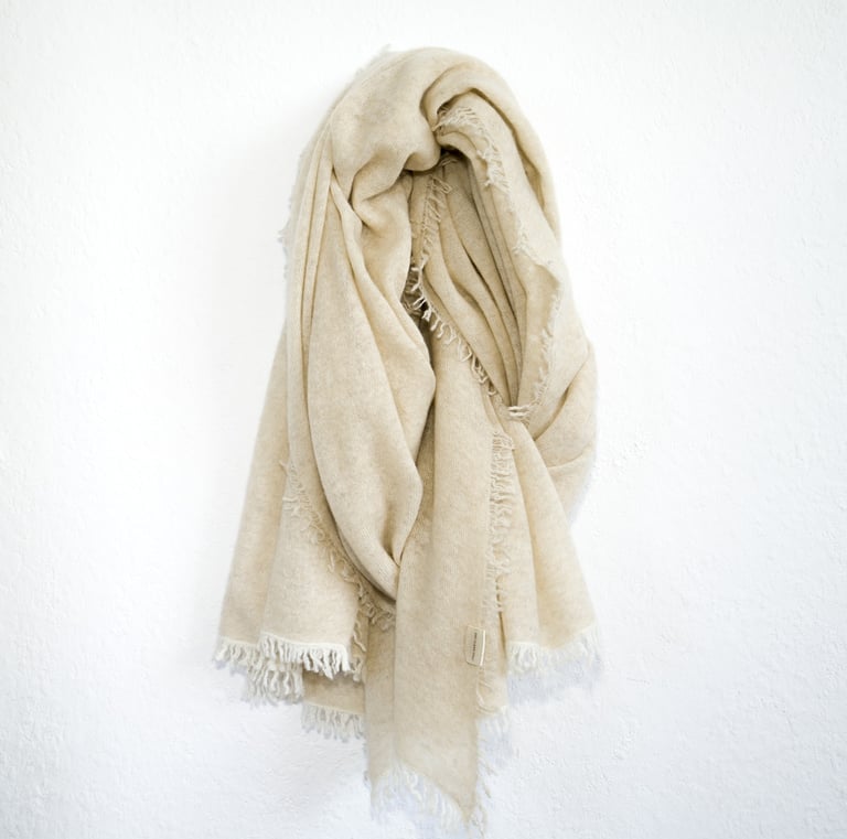 Soft cream-colored pashmina schal with fringed edges, elegantly draped on a minimal white wall.