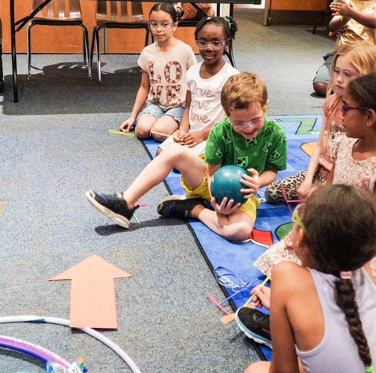 a group of children sitting on a rug in a classroom passing a weighted ball STEM