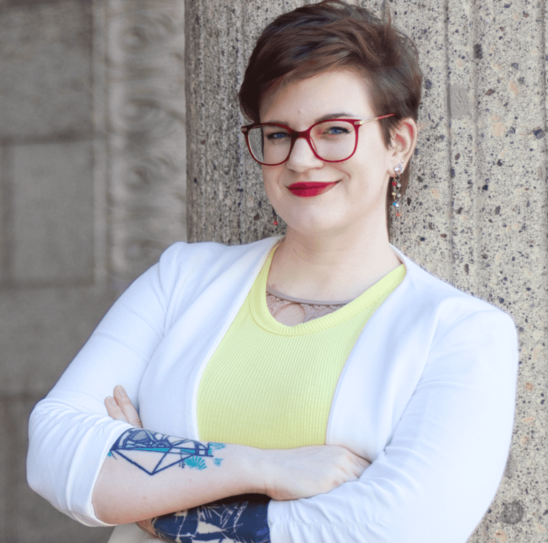 Swana Schuchmann mit Tattoos auf ihren Armen, roter Brille, gelben Top und weißem Blazer, lächelnd