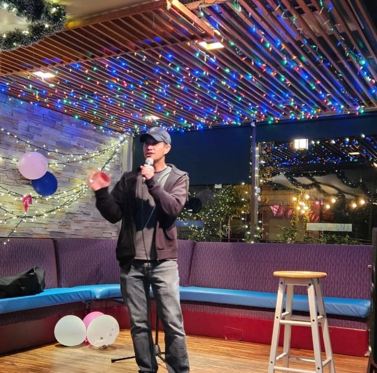 A male comedian performing stand-up comedy on a small stage with colorful festive lights and a microphone.