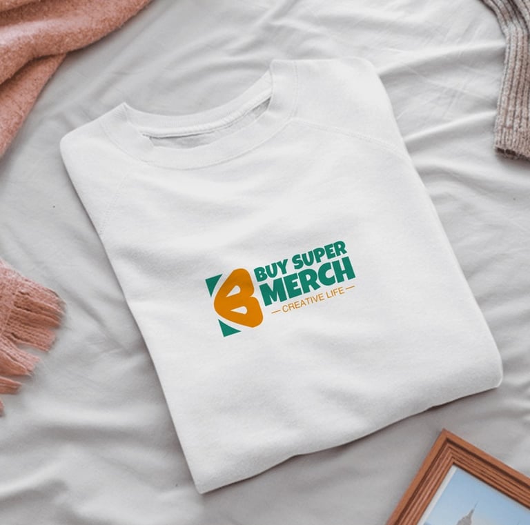 White folded sweatshirt with Buy Super Merch logo on a bed next to a pink blanket and picture frame.