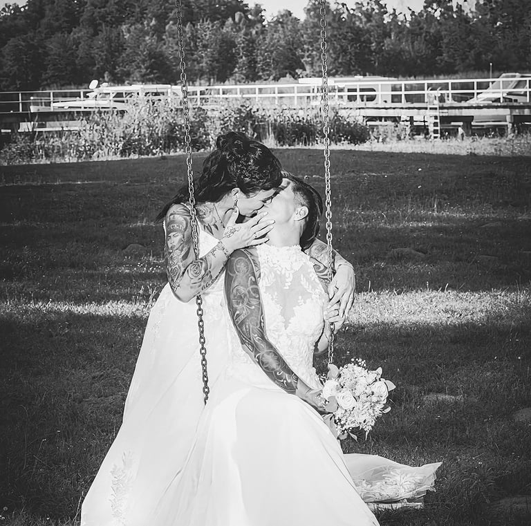  Dieses Schwarz-Weiß-Bild zeigt zwei Bräute in weißen Hochzeitskleidern, die sich küssen, während sie auf einer Schaukel sitz