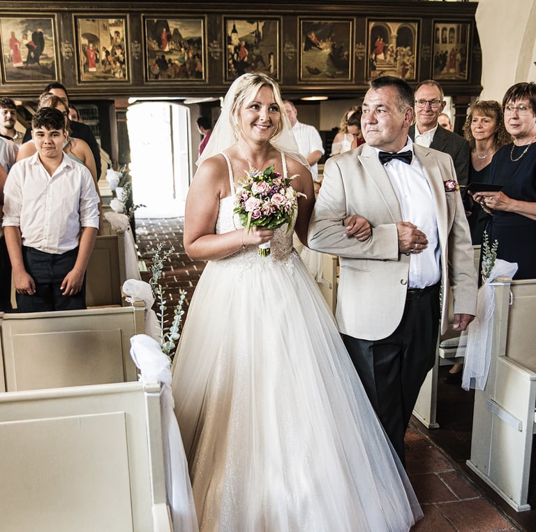  Ein Bild, das eine lächelnde Braut zeigt, die von einem Mann, wahrscheinlich ihrem Vater, den Gang einer Kirche entlanggefüh