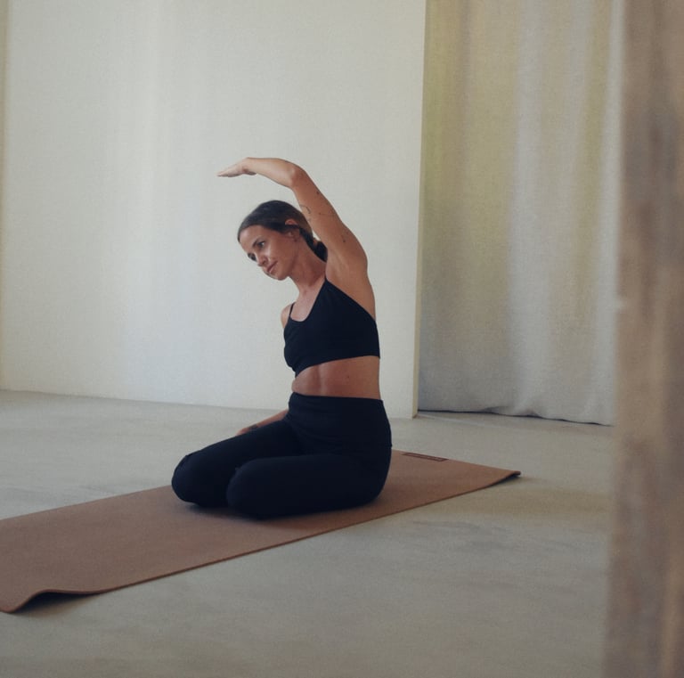 Chica sentada sobre esterilla, haciendo la postura de la sirena de Pilates. Tonos neutros y lisos.