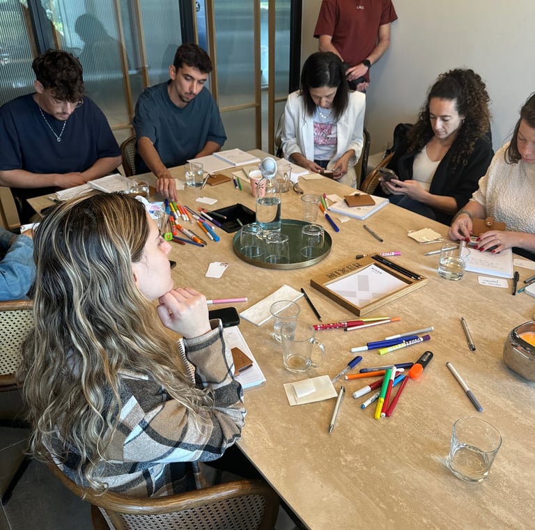 קבוצת משתתפים בסדנה יושבים סביב שולחן, עובדים על ארנקי כוחות העל שלהם עם כלי כתיבה ויצירה.