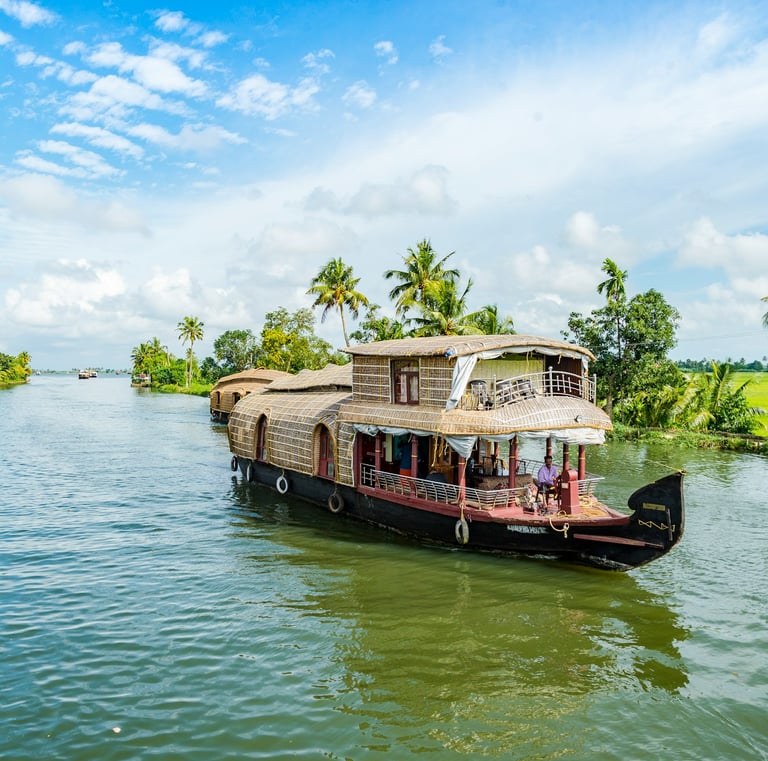 Houseboat cruising through Kerala's backwaters, surrounded by lush palm trees and vibrant greenery under a blue sky.