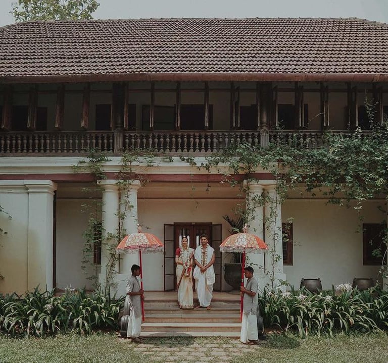 Heritage stay in Kerala featuring a couple in traditional attire, flanked by attendants holding umbrellas.