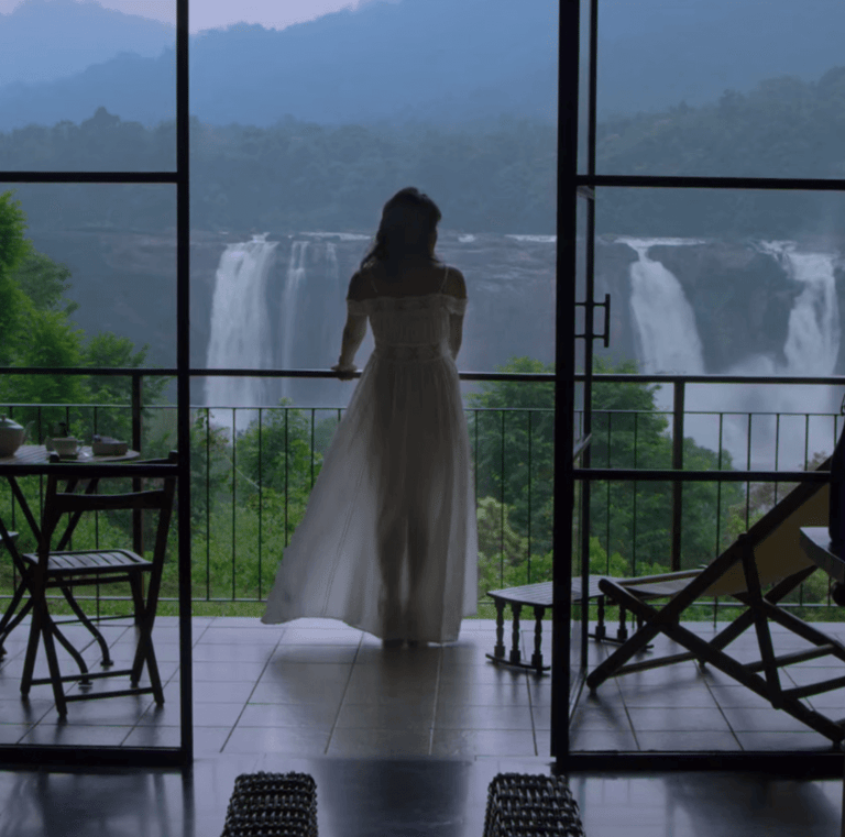 Woman in white dress standing on balcony overlooking Athirappilly Falls, framed by glass doors.