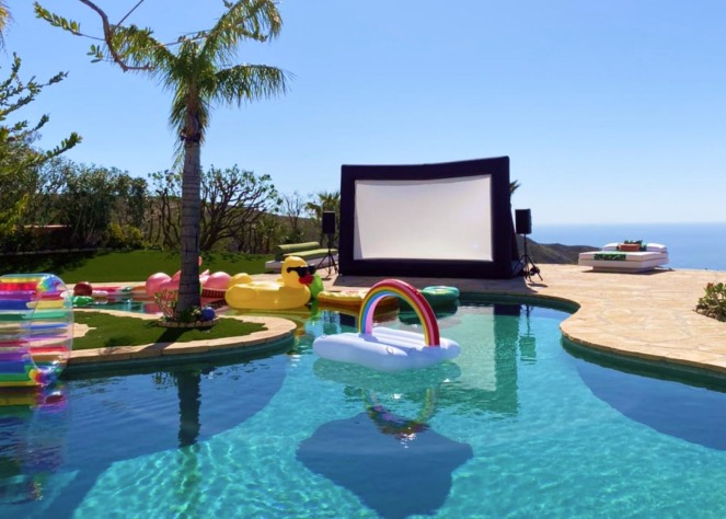 A swimming pool is surrounded by inflatable toys under a clear sky, with a large outdoor screen rental and speakers set 
