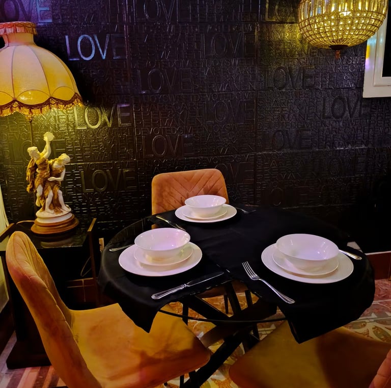 Elegant dining table set for three with white bowls on a black cloth, featuring a textured LOVE wall and vintage lamp.