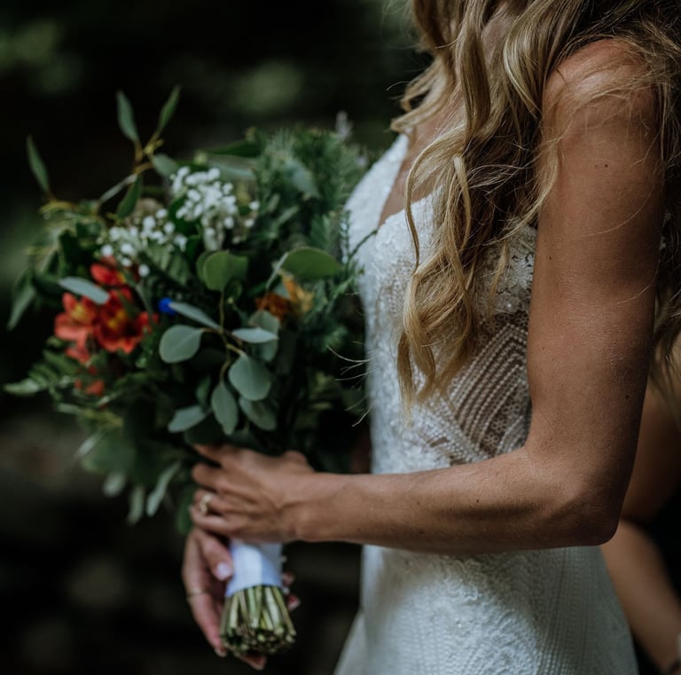 A smiling bride in a lace wedding dress holding a rustic wildflower bouquet with greenery.