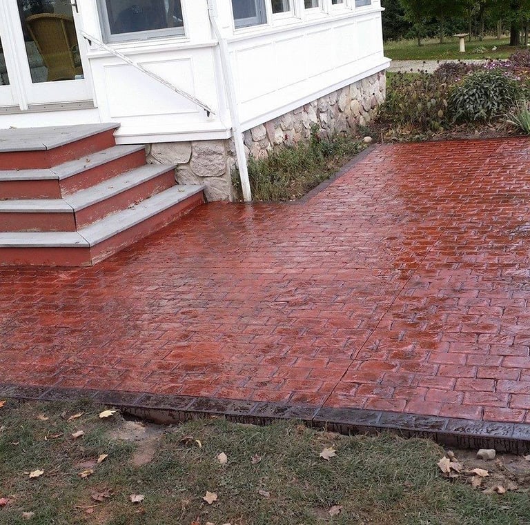Red stamped concrete patio with a decorative brick pattern installed next to a house's wooden stairs.