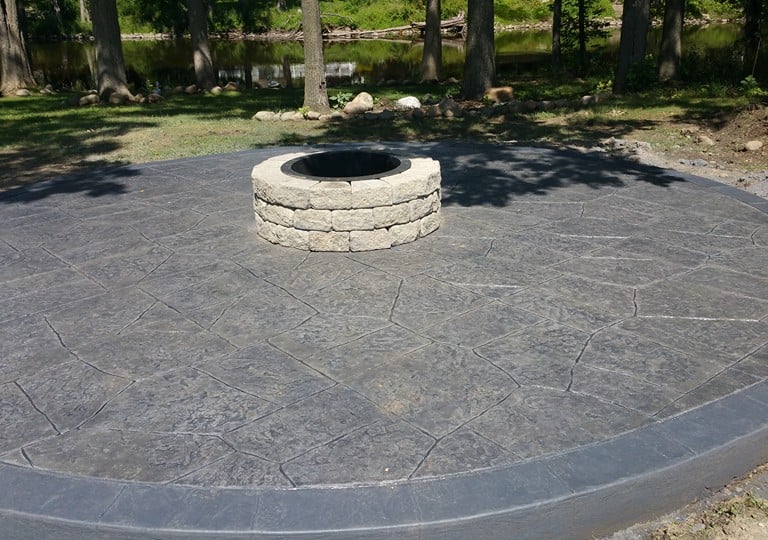 Circular stamped concrete patio with a stone fire pit near a backyard river.