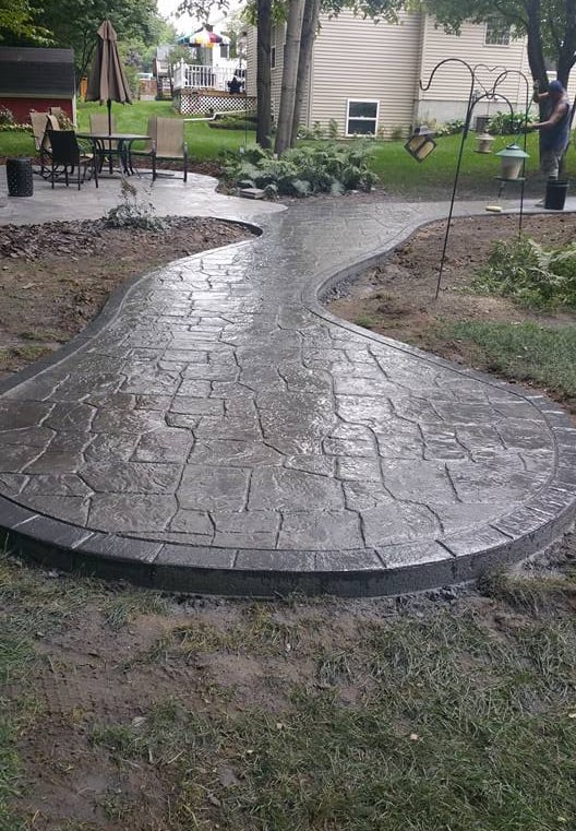 Curved dark gray stamped concrete walkway and patio in a residential backyard landscape.