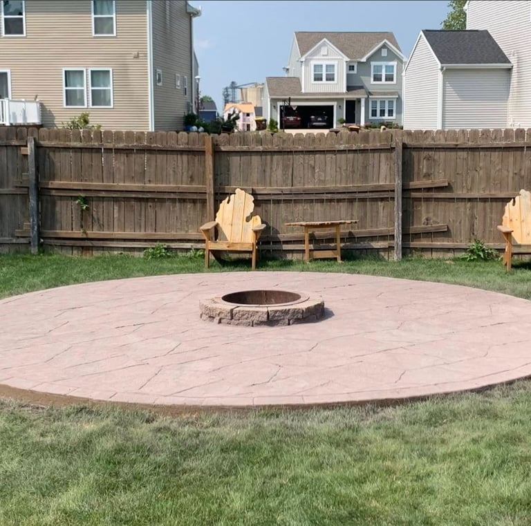 Circular stamped concrete patio with central fire pit and Adirondack chairs in a fenced backyard.