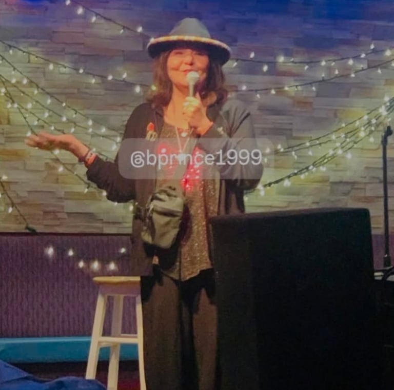 Female stand-up comedian performing on a stage with string lights and a stone wall background.