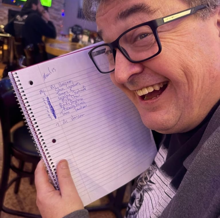 Smiling man in glasses holding a handwritten list in a notebook at a dimly lit bar.