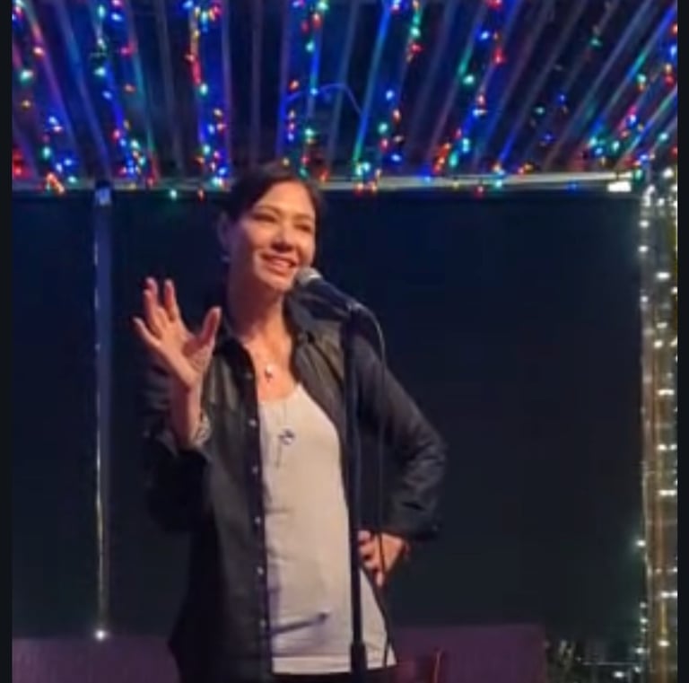 Female stand-up comedian performing on a stage under colorful string lights at a comedy club.