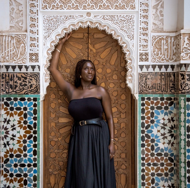 A woman in a black dress posing before an ornate Moroccan door with intricate tilework and carvings.