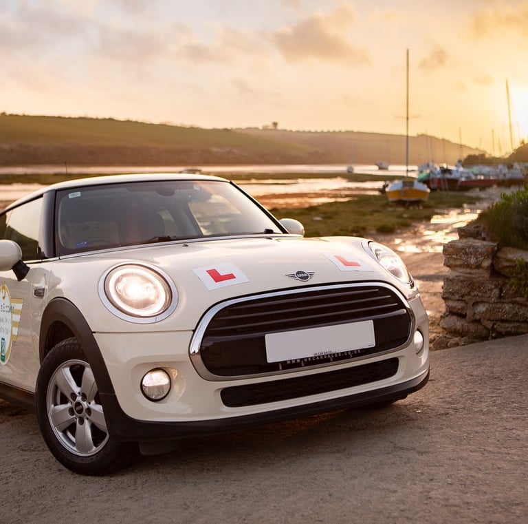 White Mini Cooper driving school car with L-plates parked at a coastal harbor at sunset.