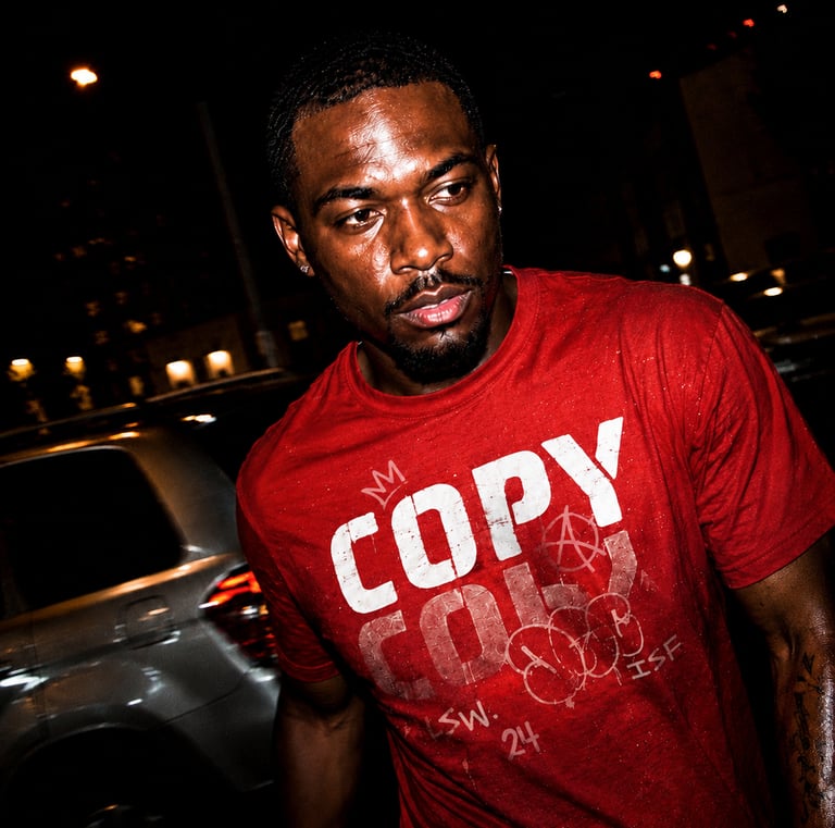 A dark, intense portrait of a man in a red "COPY" t-shirt, sweat glistening on his face, in a nighttime urban setting.