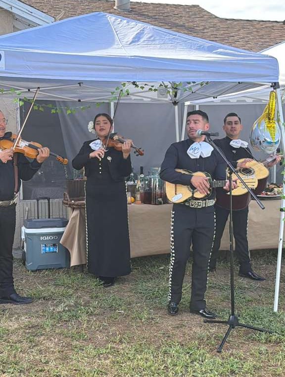 A traditional mariachi band in black charro suits performing with violins and guitars under a white canopy.