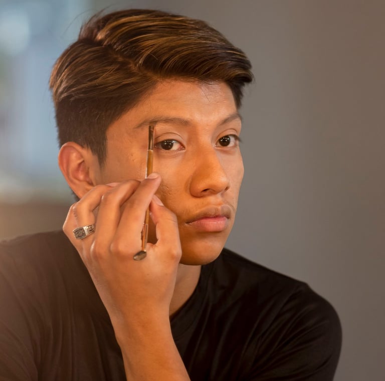 Un joven se aplica maquillaje de cejas con una herramienta de pincel profesional frente a un espejo.