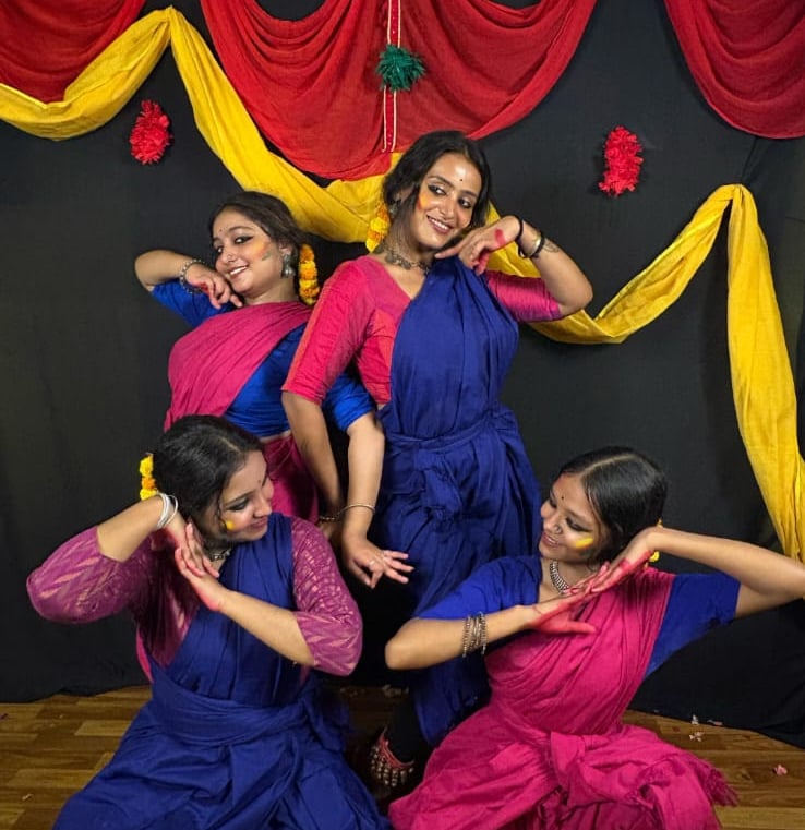 Four Indian women in traditional blue and pink sarees performing a folk dance on a decorated stage.