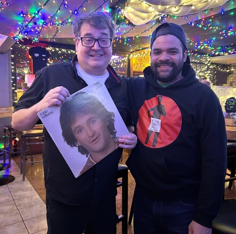 Two men smiling in a restaurant decorated with holiday lights while holding a Robin Williams vinyl record.