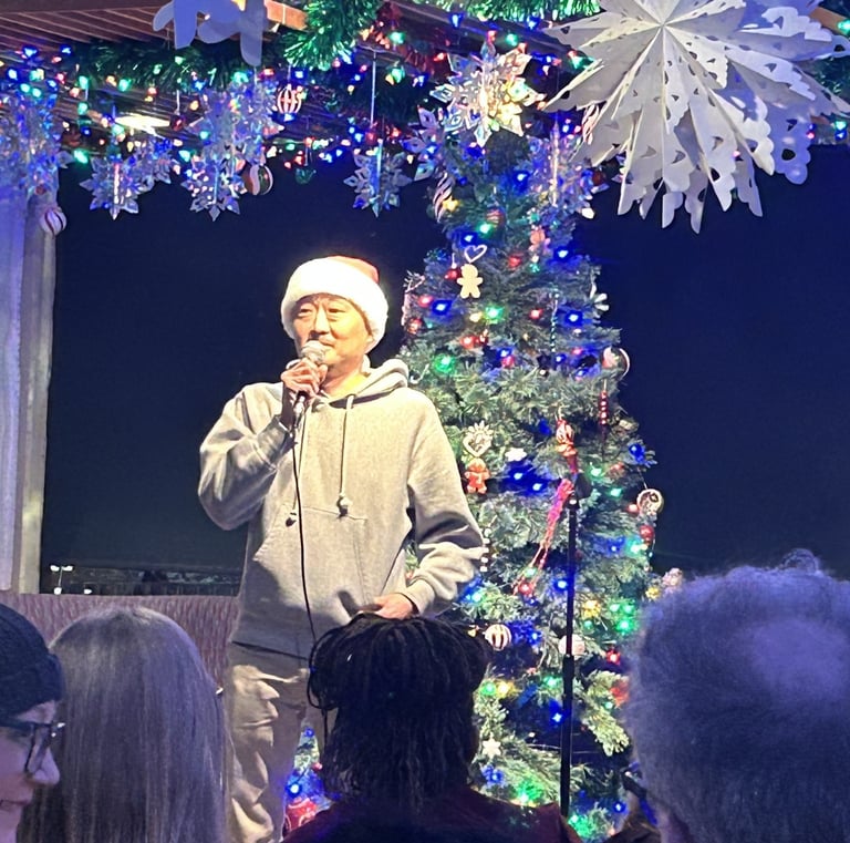 A man in a Santa hat performing holiday karaoke beside a lighted Christmas tree and festive decorations.