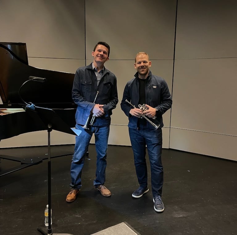 George Chase and Caleb Hudson smile, holding piccolo trumpets, during a rehearsal break.