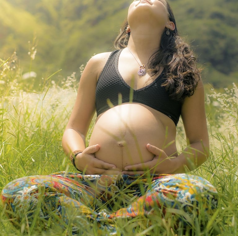 a young pregnant woman wearing a crop top is sitting in a grass field and looking up to the sky while holding her belly