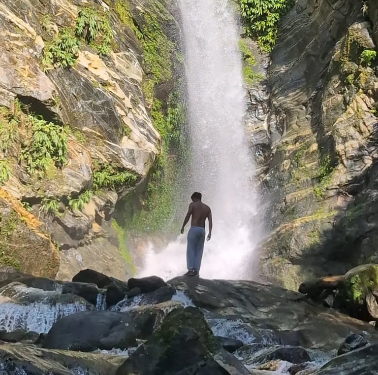 A man stands on rocks looking at a powerful tropical waterfall cascading down a lush cliffside.