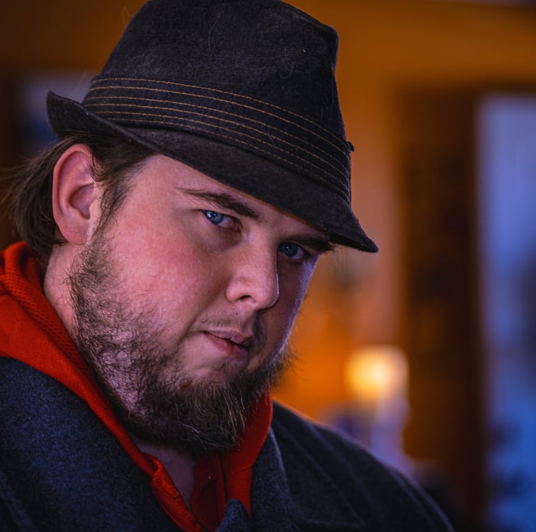 A man with a beard wearing a dark fedora and jacket in a close-up portrait in soft lighting. Athens,