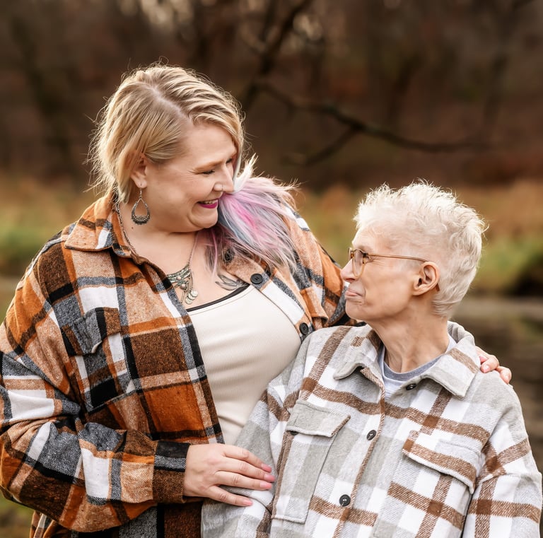 A blonde woman with pink hair highlights and an older woman wearing flannel shirts smiling outdoors.