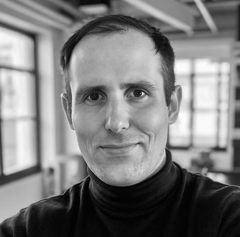 Black and white professional headshot of a smiling man wearing a black turtleneck in a bright studio office.