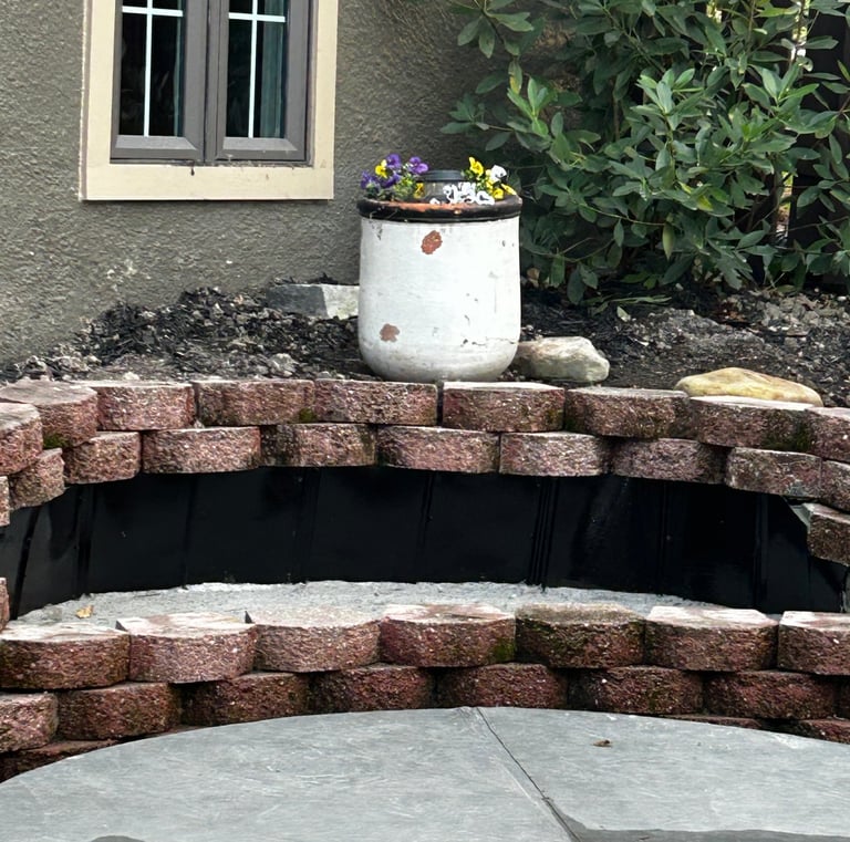 Curved fire pit retaining wall made of red stone blocks next to a white flower pot and house window.