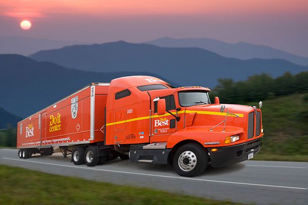 An orange Do it Best semi-truck driving on a mountain highway at sunset.