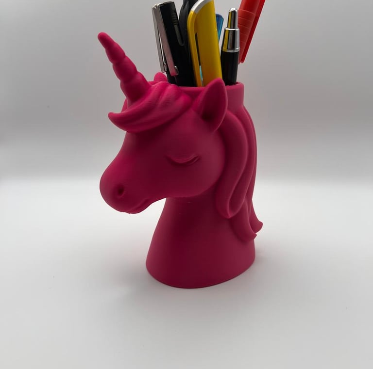 A pink unicorn head pencil holder filled with colorful pens and office supplies on a white background.