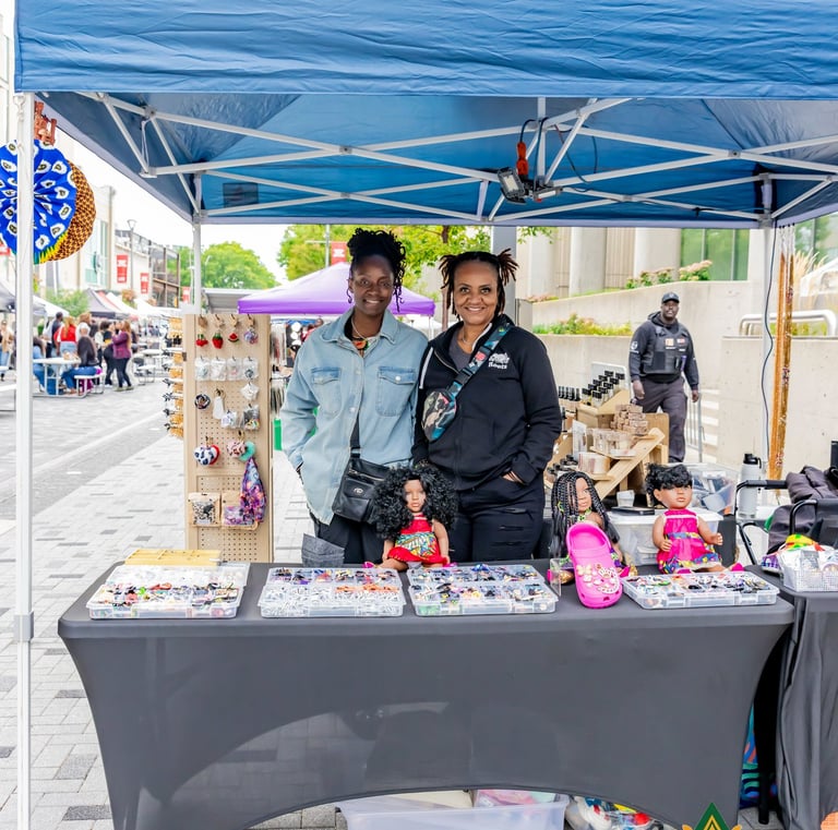 Two smiling women standing at their craft stall under a blue canopy at the Afro Fiesta Festival.