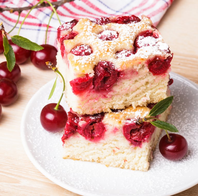 A stack of two cherry cake bars dusted with powdered sugar on a white plate, garnished with fresh whole cherries.