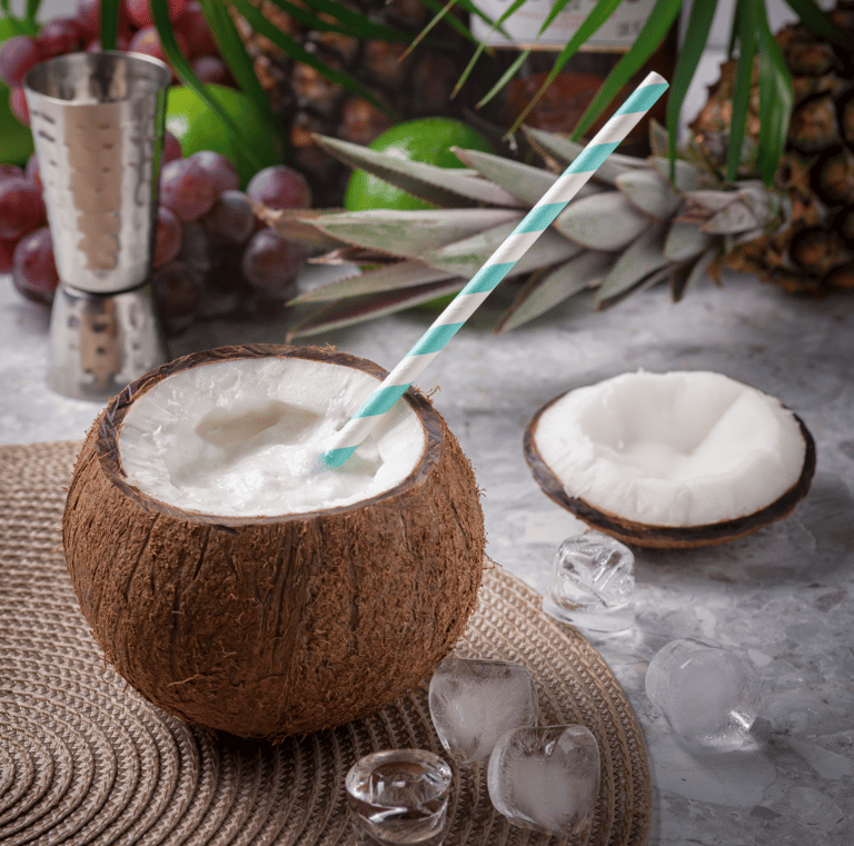 A tropical coconut drink with a striped straw, served in a coconut shell with ice cubes and pineapple nearby.