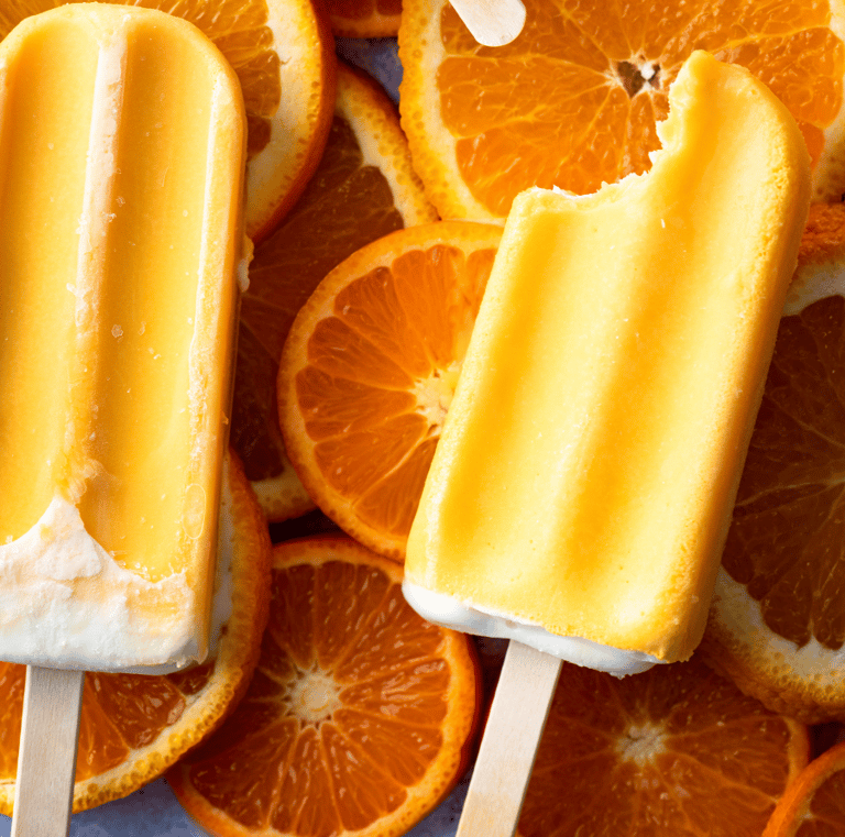 Two orange cream popsicles, one with a bite taken out, resting on a background of fresh, juicy orange slices.