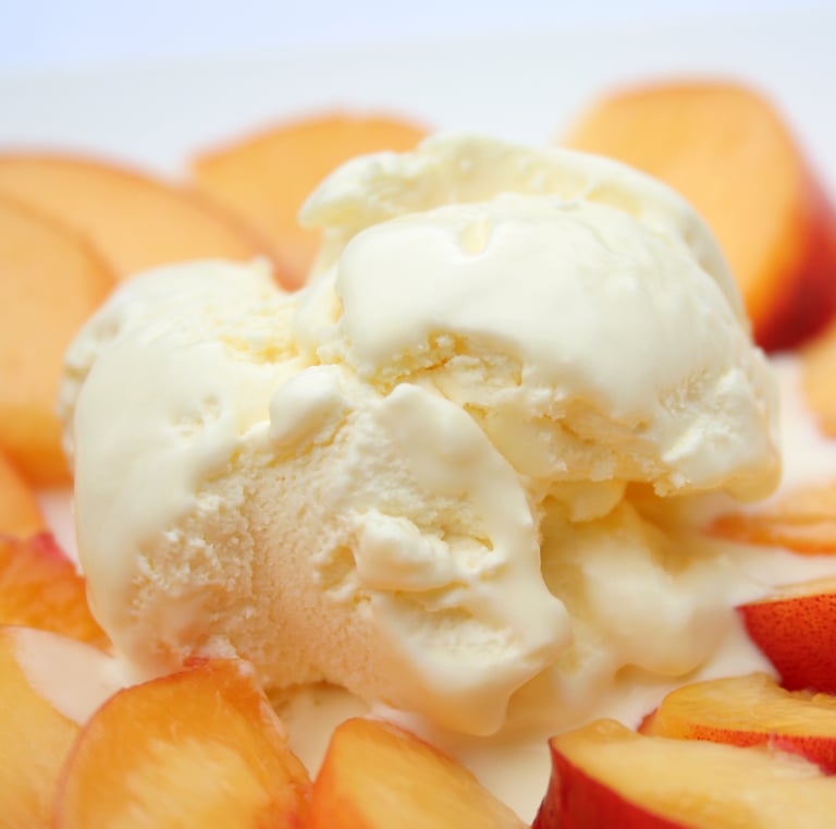 Close-up scoop of creamy vanilla ice cream melting on top of a bed of fresh, sliced yellow peaches on a white plate.