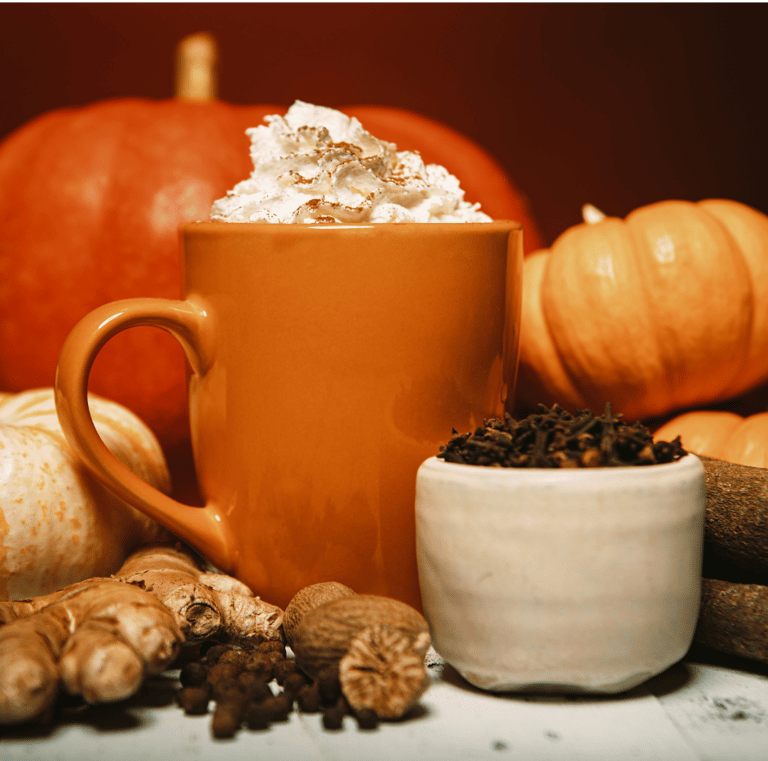 An orange mug of hot pumpkin spice drink topped with whipped cream, surrounded by whole pumpkins, ginger, and spices.