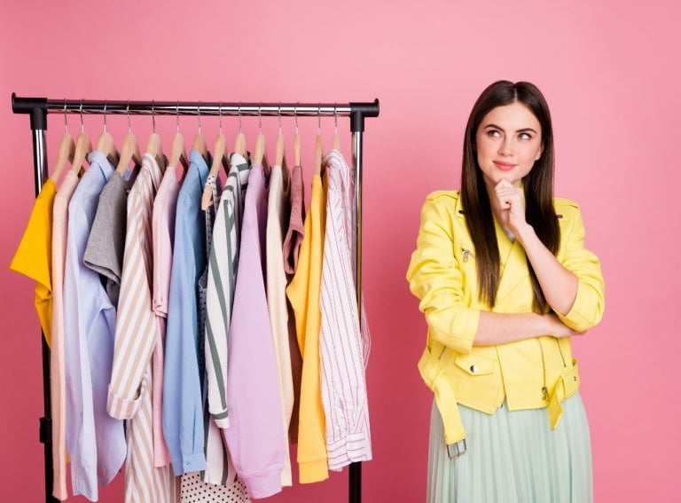 Brunette in colorful clothes looking thoughtful near hanging shirts