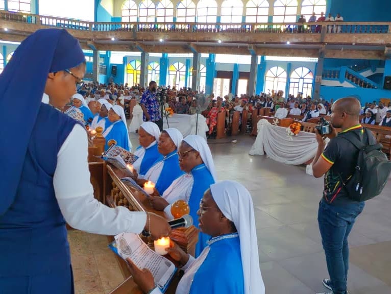 Catholic nuns in blue habits holding lit candles during a church ceremony with a congregation in the background.