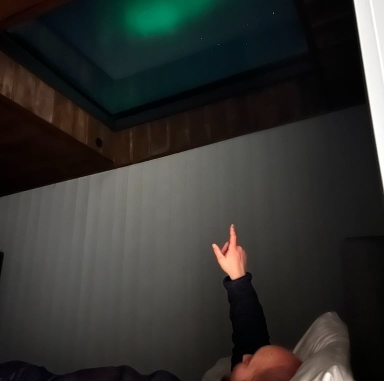 A person lying in bed points up at the glowing green Northern Lights visible through a glass ceiling skylight.