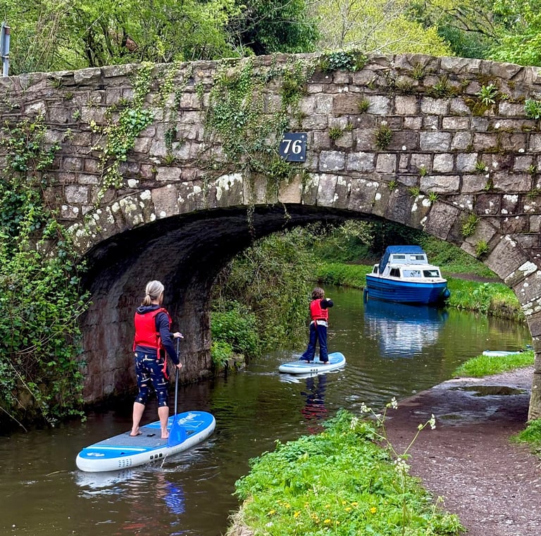 A family paddleboard under a stone bridge on the Monmouthshire and Brecon canal in the Brecon Beacons, surrounded by nature.